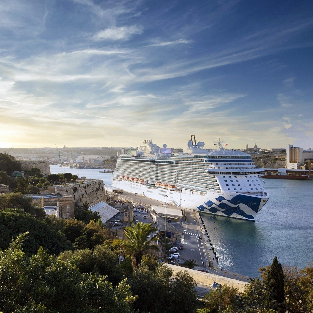 Aerial view of a white and blue Princess cruise ship docked at a harbor with stone fortifications, lush greenery, and a Mediterranean cityscape visible in the background under a bright sky at sunset.