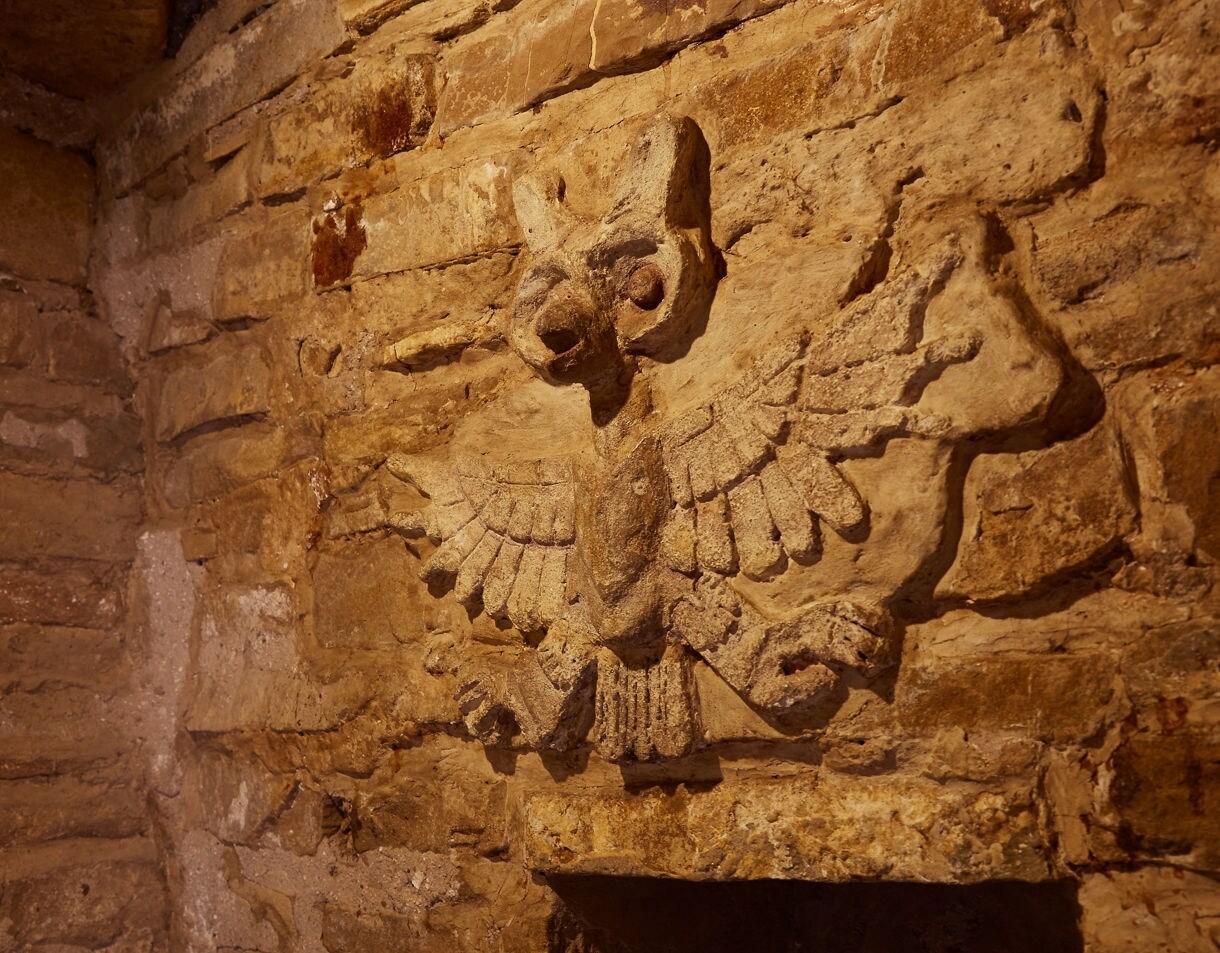 Stone-carved owl relief on the wall of a Zapotec tomb, etched into weathered brown stone blocks.