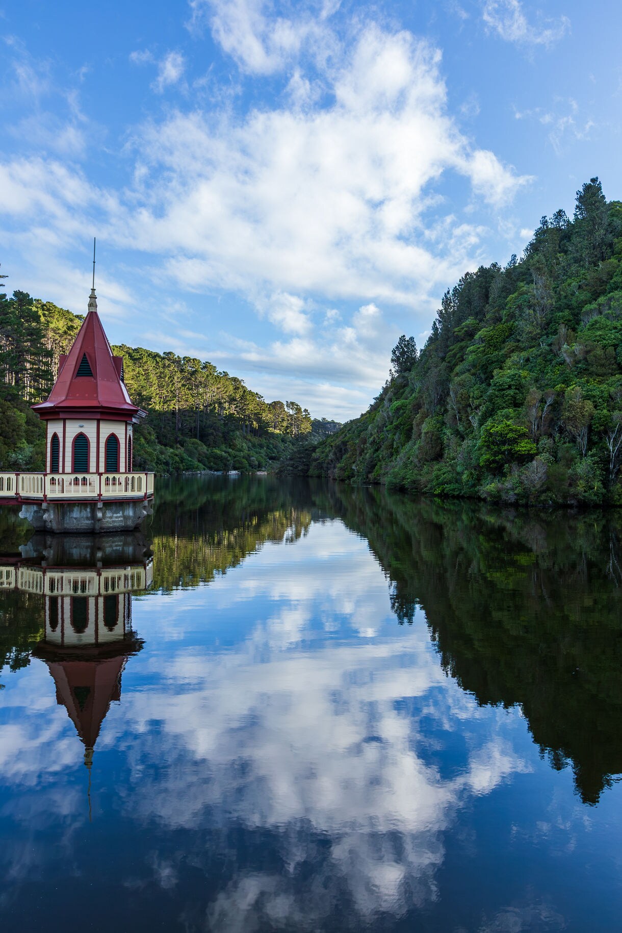 Red-roofed pavilion beside a calm lake at Zealandia wildlife sanctuary, reflecting trees and clouds in still water.