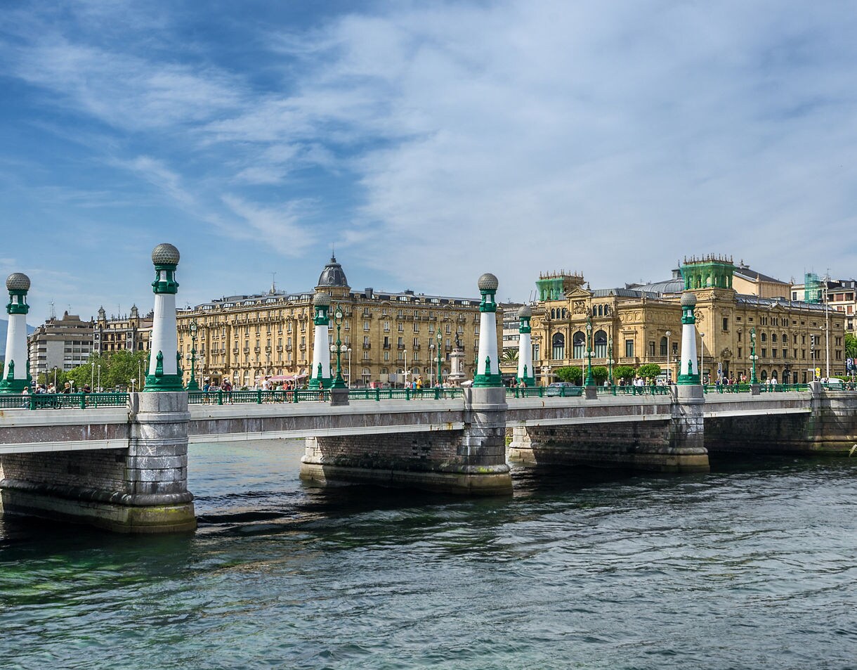 The Zurriola Bridge in San Sebastián featuring white and green Art Deco lampposts, spanning the Urumea River with historic sandstone buildings in the background.