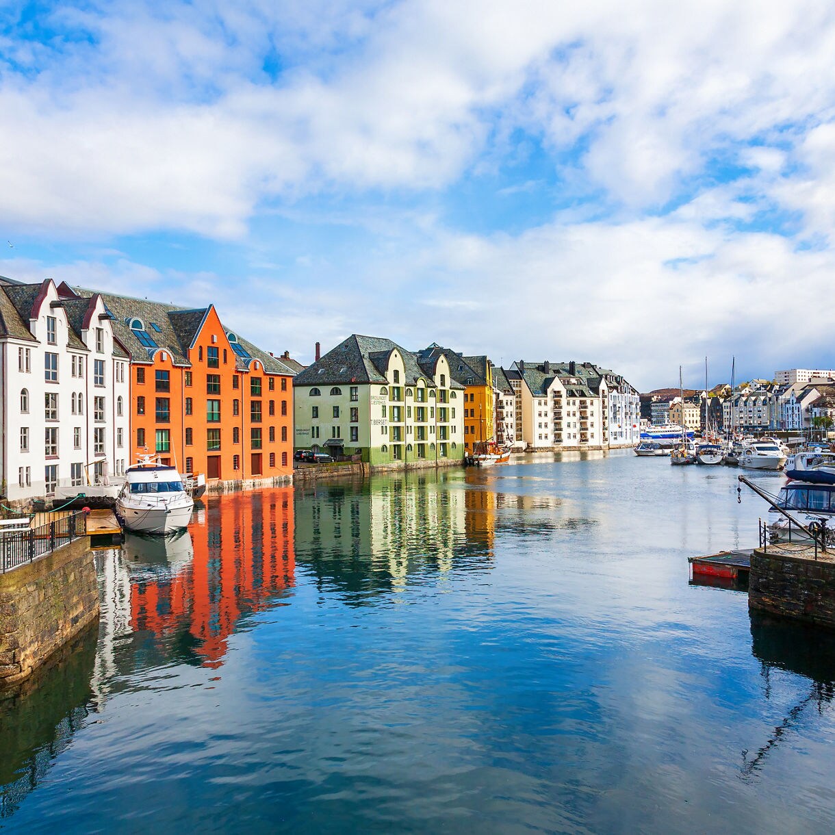Colorful Art Nouveau buildings line a calm canal in Ålesund with boats moored along the water under a partly cloudy sky.